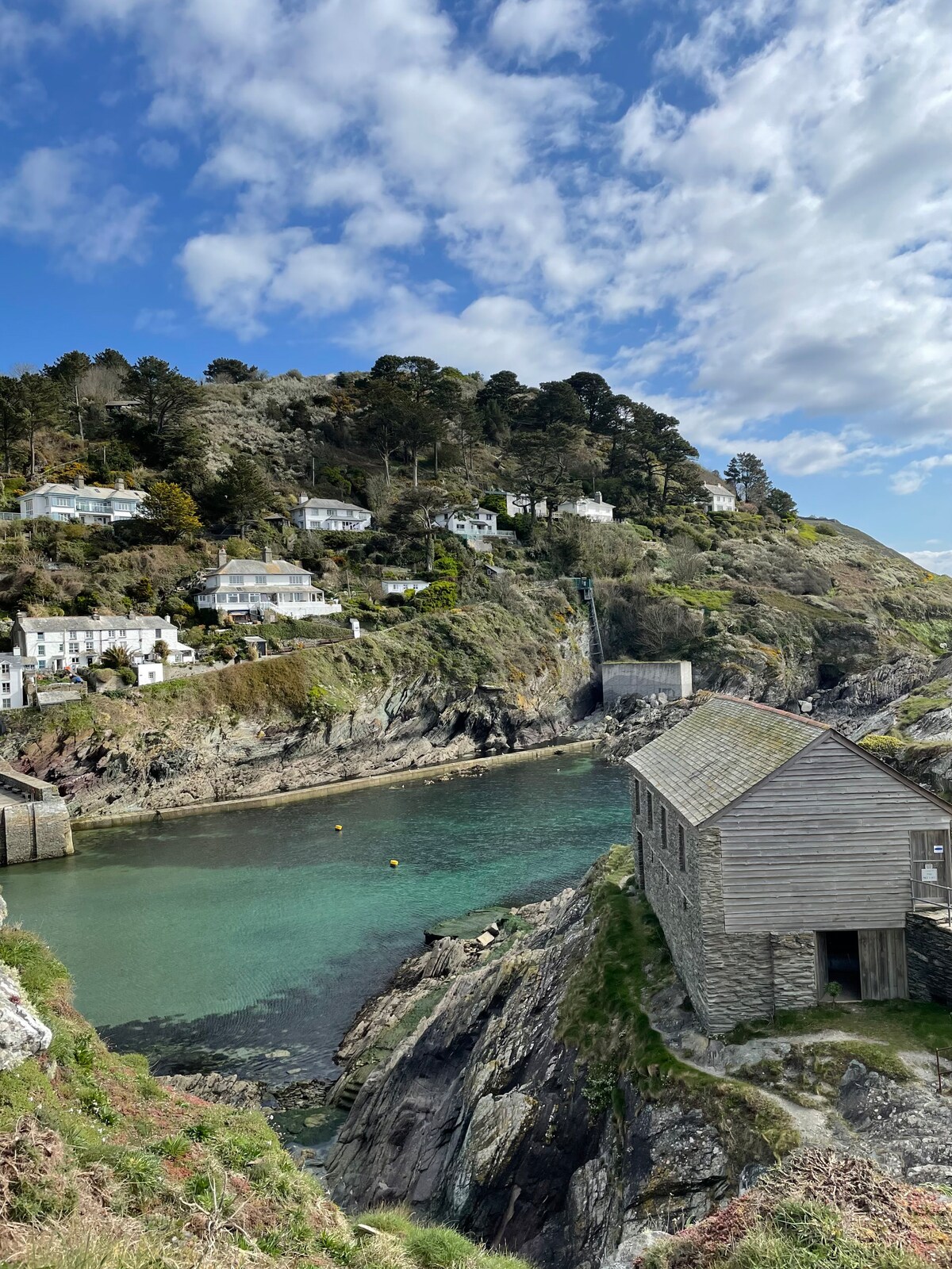 A scenic view of a rocky coastline features a calm inlet with clear water. Houses are situated on the hillside, surrounded by trees. A small, rustic structure can be seen at the water's edge, with the landscape embellished by scattered clouds in a blue sky.