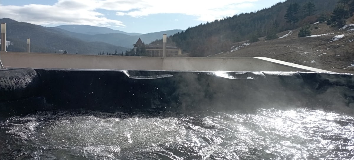 An outdoor jacuzzi filled with steaming mineral water is depicted, surrounded by scenic mountains and greenery. The blue sky is visible overhead, accentuating the tranquil environment. The water reflects sunlight, creating a calming atmosphere suitable for relaxation.