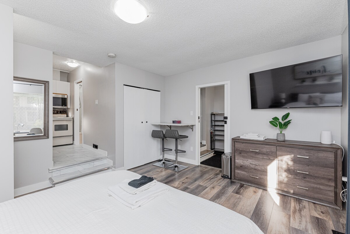 A spacious Bachelor Suite is captured, featuring a crisp white bedding set on a generous bed. A smart TV is mounted on the wall across from a compact kitchenette and dining area. Natural light enters through a nearby window, illuminating the practical furniture arrangement.
