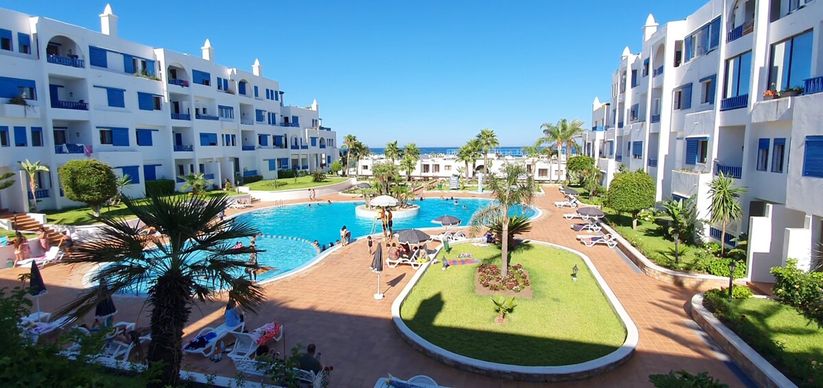 A serene outdoor area is depicted, featuring landscaped gardens and a central pool. Sun loungers are positioned around the pool, with umbrellas providing shade. The backdrop includes multiple white buildings, and the sea is visible in the distance under a clear blue sky.