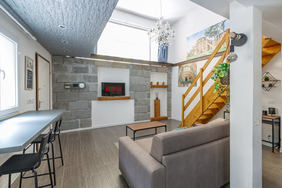 An open-concept living area features high ceilings and large windows. A comfortable gray sofa faces a wall-mounted Smart TV displaying the Netflix logo. A wooden staircase leads to a loft area, while a long black dining table with high chairs occupies one side of the space.