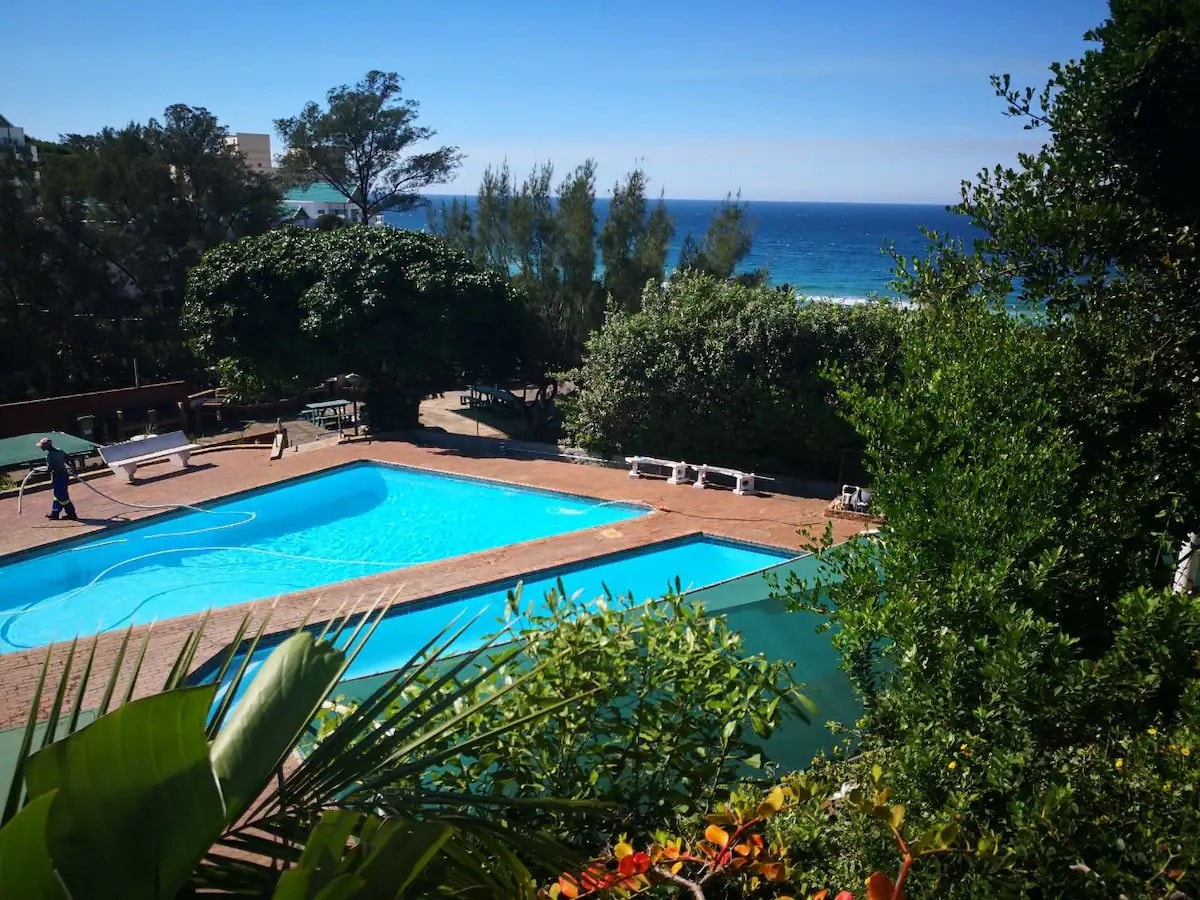 A sparkling swimming pool is framed by vibrant greenery, with lounge chairs arranged nearby. The sea can be seen in the background, creating a scenic coastal view. Sunlight reflects off the water, enhancing the inviting atmosphere of the outdoor space.