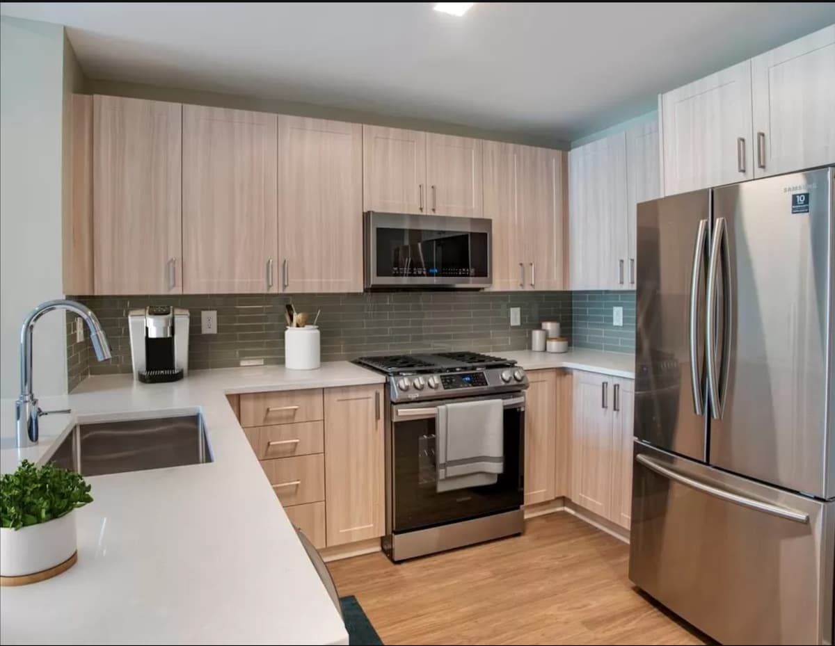 A modern kitchen features light wood cabinetry and stainless steel appliances, including a large refrigerator and a gas stove. Countertops are made of smooth, white material, complemented by a grey tiled backsplash. The workspace is well-lit and includes various kitchen utensils organized neatly.