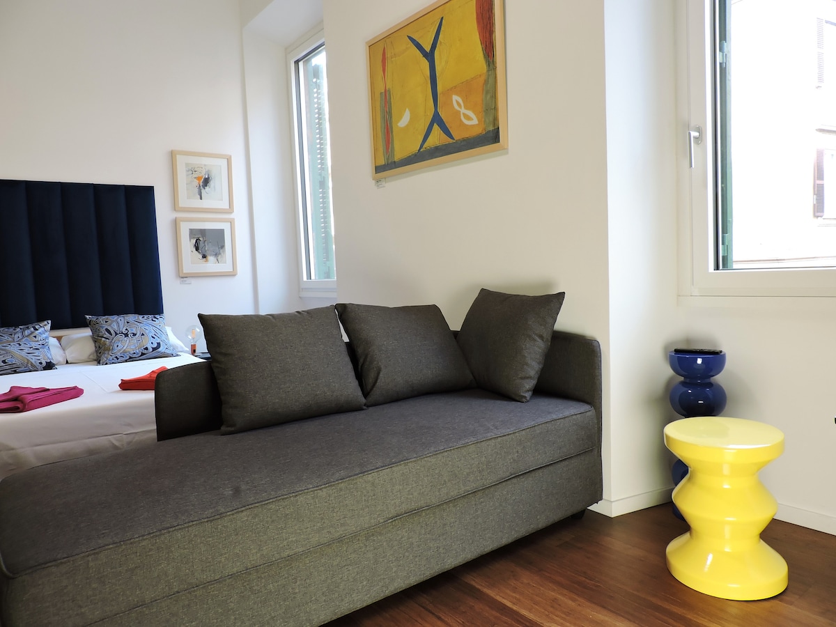 A comfortable gray sofa is positioned beside a bright yellow side table. An abstract artwork hangs above, complementing the modern design. Natural light enters through a window adorned with light curtains, illuminating the cozy seating area adjacent to the bed.