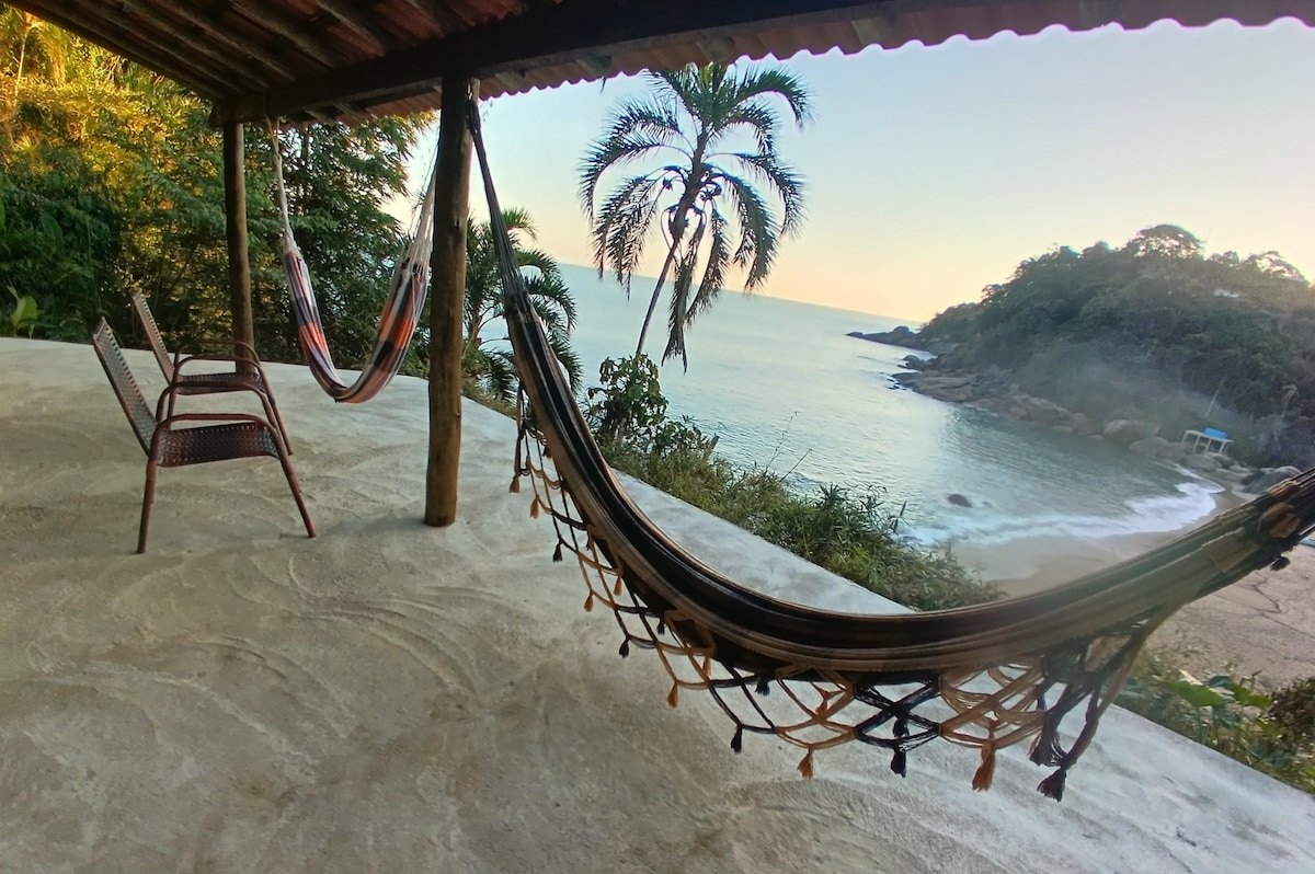 A serene outdoor space features two hammocks hanging from wooden supports, inviting relaxation while overlooking the ocean. A small table and a chair are placed nearby, and lush greenery flanks the area. The view includes gentle waves and a distant coastline, illuminated by soft morning light.