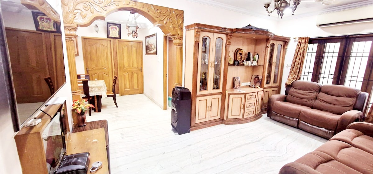 The living area features a traditional wooden arch and cabinets, offering a blend of elegance and comfort. A cozy sofa set is positioned to provide ample seating, and a television stands against one wall. Natural light filters in through large windows, enhancing the inviting space.