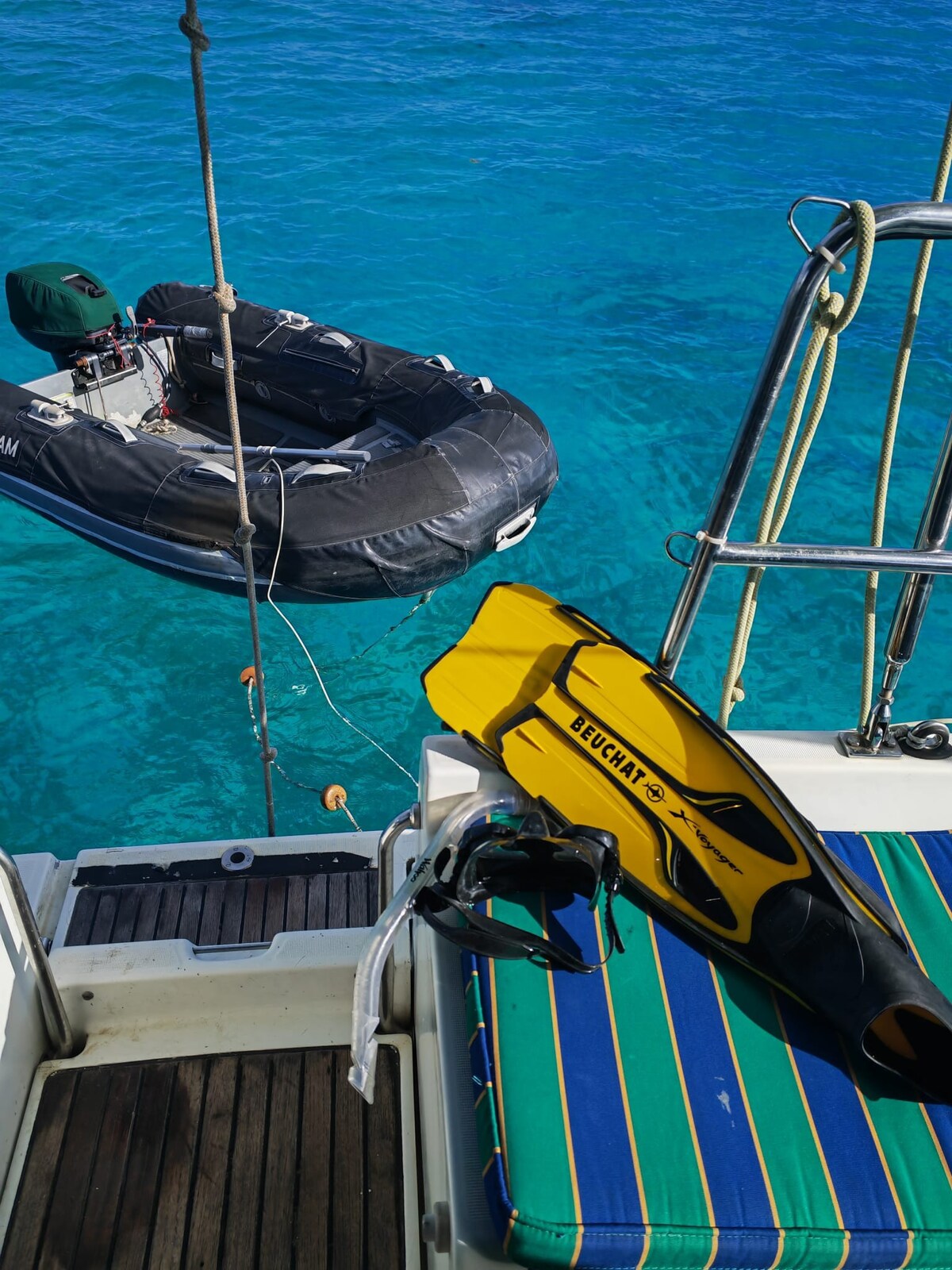A small inflatable boat hangs from the side of a larger vessel, gently rocking above clear, turquoise water. A pair of yellow snorkeling fins and a mask rest on a striped cushion, ready for exploration. The wooden deck offers a natural look contrasting with the vibrant blue below.
