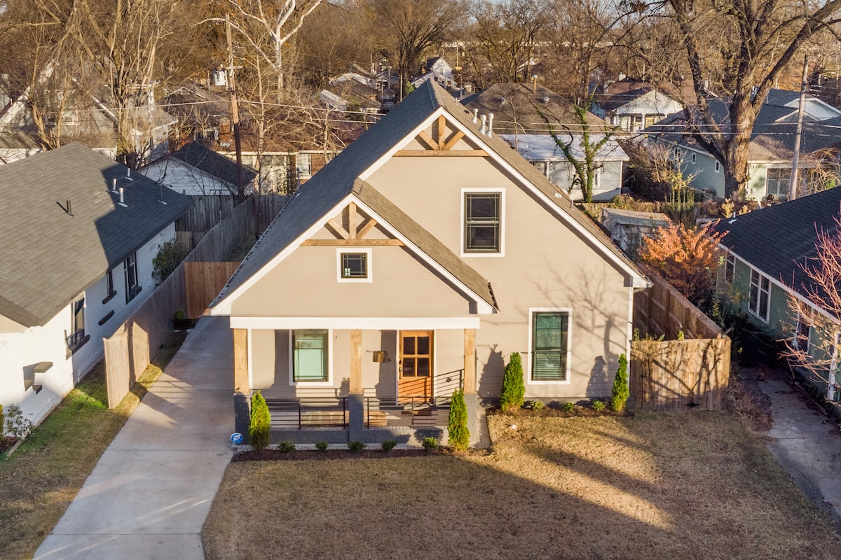 The property features a two-story house with a light-colored exterior, designed with architectural details such as exposed wood beams. A spacious front yard is bordered by low shrubs, while a paved driveway leads to the entrance, providing access to the home.