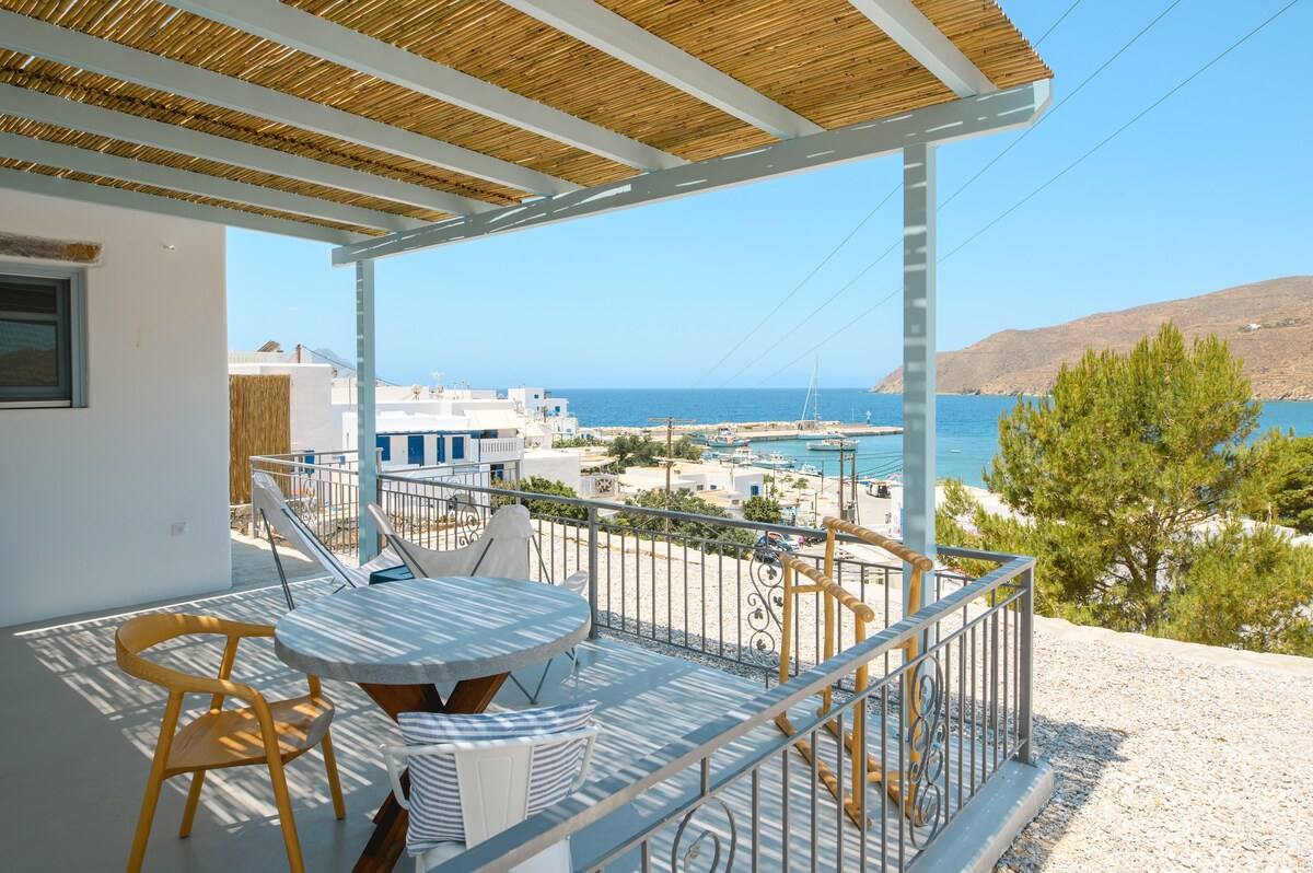 A spacious outdoor terrace is featured, equipped with a round table and several chairs. The area provides unobstructed views of the sea and surrounding landscape. A striped hammock hangs for relaxation. The structure is shaded by a straw canopy, enhancing comfort during sunny days.
