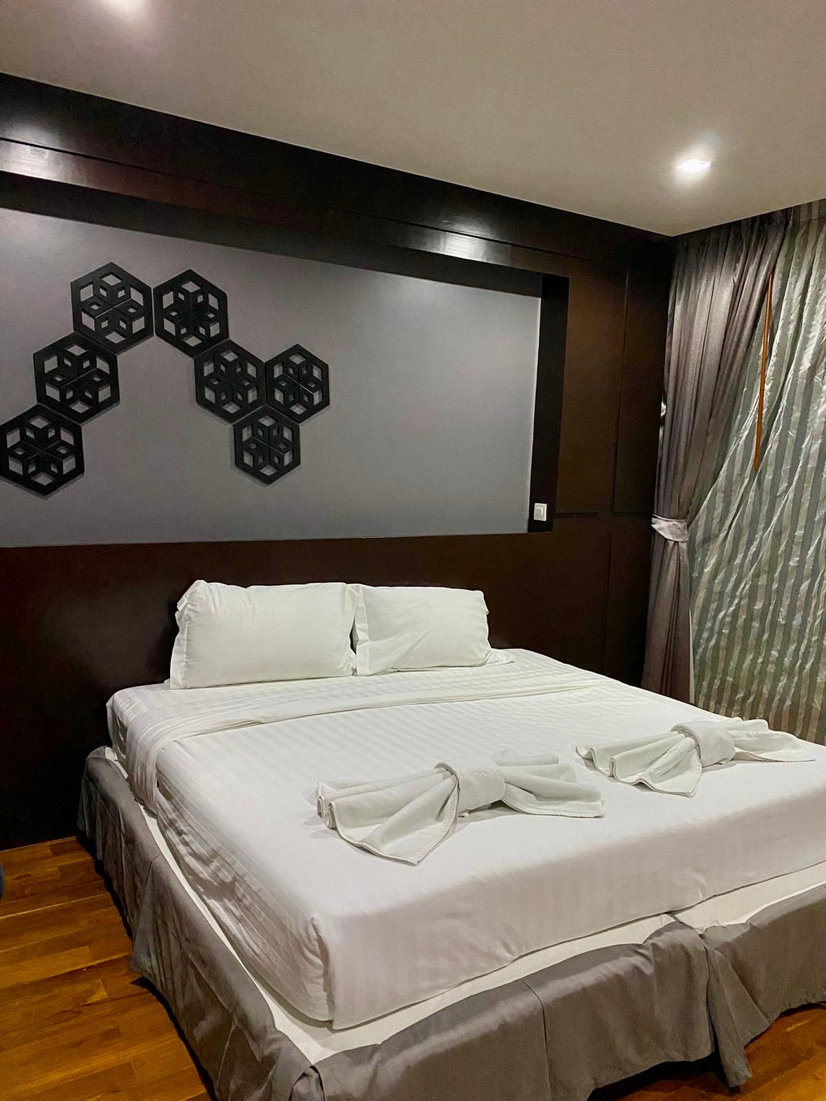 A spacious sleeping area features a neatly made bed with white linens. Two decorative towels are arranged on the bed, and a geometric wall art piece is positioned above the headboard. Soft, natural light enters through sheer curtains, enhancing the serene atmosphere.