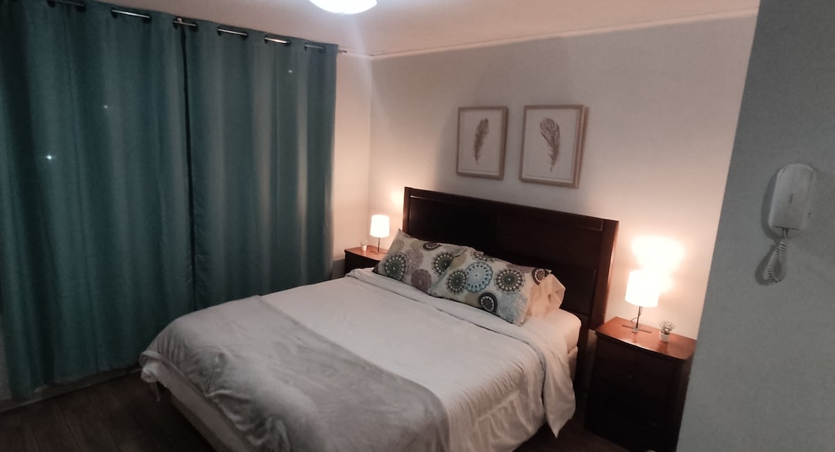 A well-lit sleeping area features a spacious bed dressed in neutral linens. Two bedside tables are positioned on either side, each topped with a lamp. Light blue curtains frame the window, while decorative wall art completes the calming atmosphere.