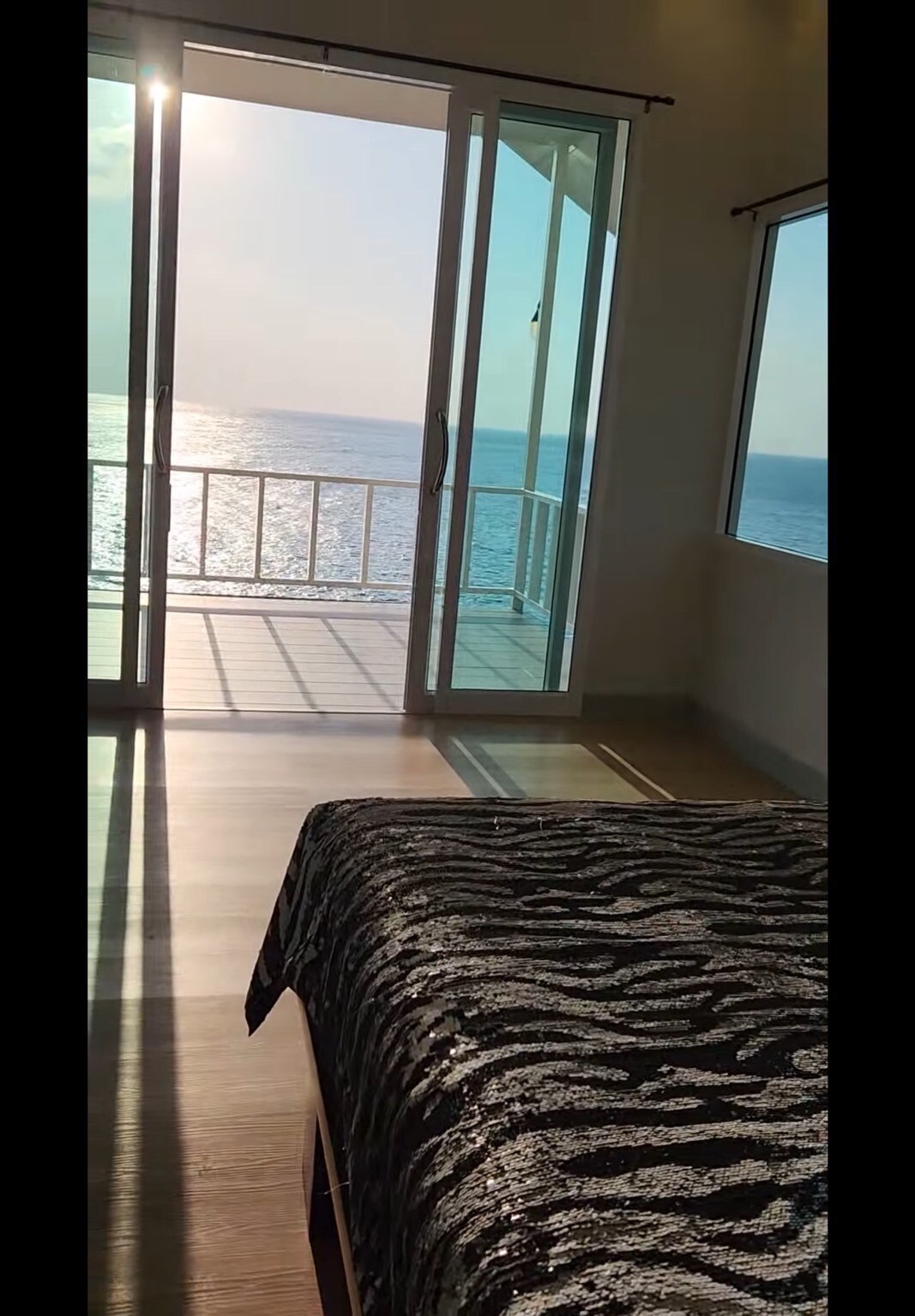 A serene bedroom features a spacious bed covered with textured black and white bedding. Large glass doors open to a balcony overlooking the ocean, allowing natural light to fill the room. Views of sparkling water can be glimpsed through the windows.