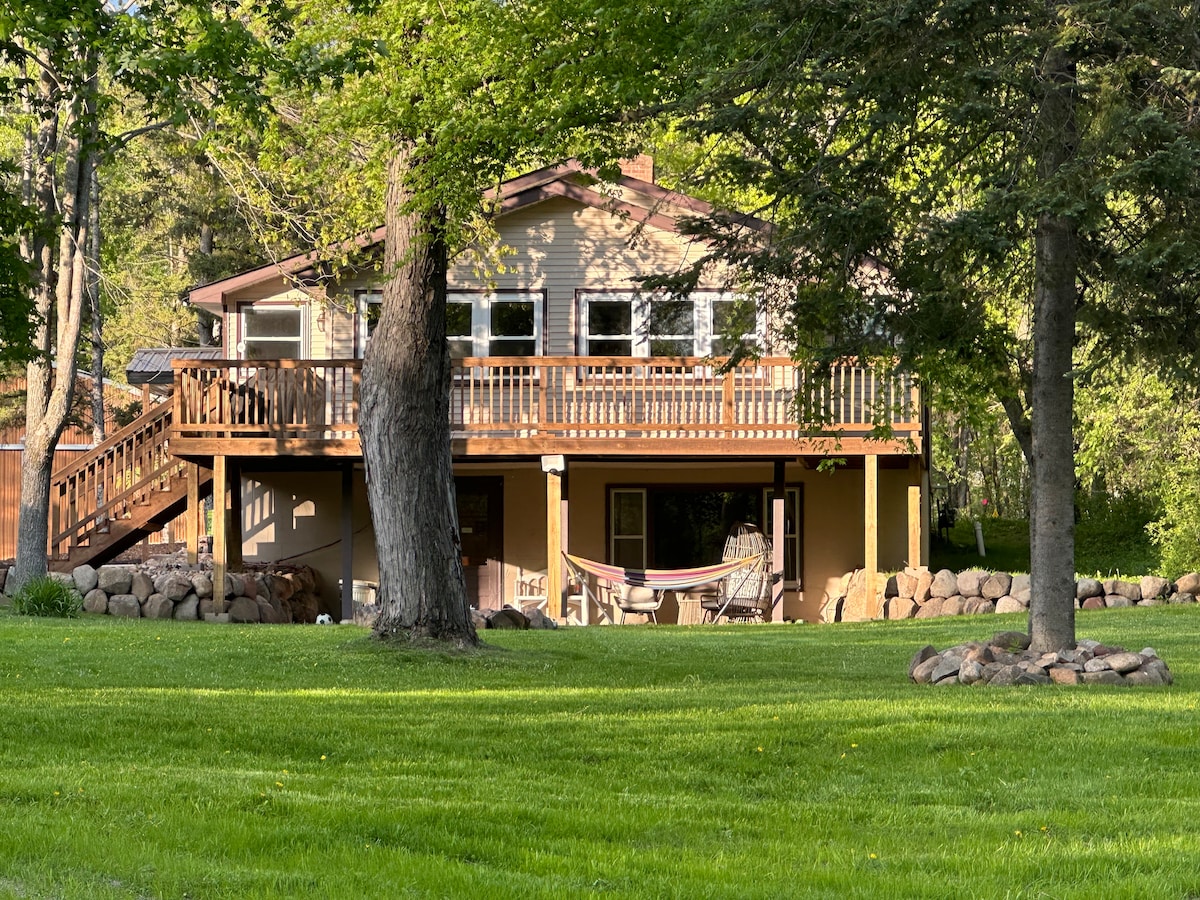 A two-story house is nestled among trees, featuring a large deck with wooden railings. A hammock hangs near the ground level, surrounded by well-maintained grass and smooth stones. The exterior showcases warm tones and multiple windows, inviting natural light into the living spaces.
