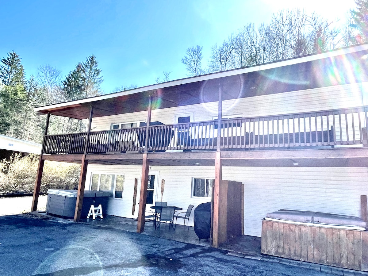 The exterior of a ground-level duplex cabin is shown, featuring wooden pillars supporting a spacious balcony. A hot tub and a wooden storage area are visible, alongside outdoor seating. The setting is semi-wooded with trees in the background and sunlight filtering through.
