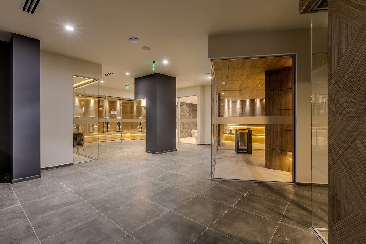 A spacious lobby area showcases modern design with large glass walls that allow natural light to fill the space. The gray tiled floor complements the warm wooden accents on the wall. Thoughtful lighting highlights various seating and relaxation areas throughout the room.