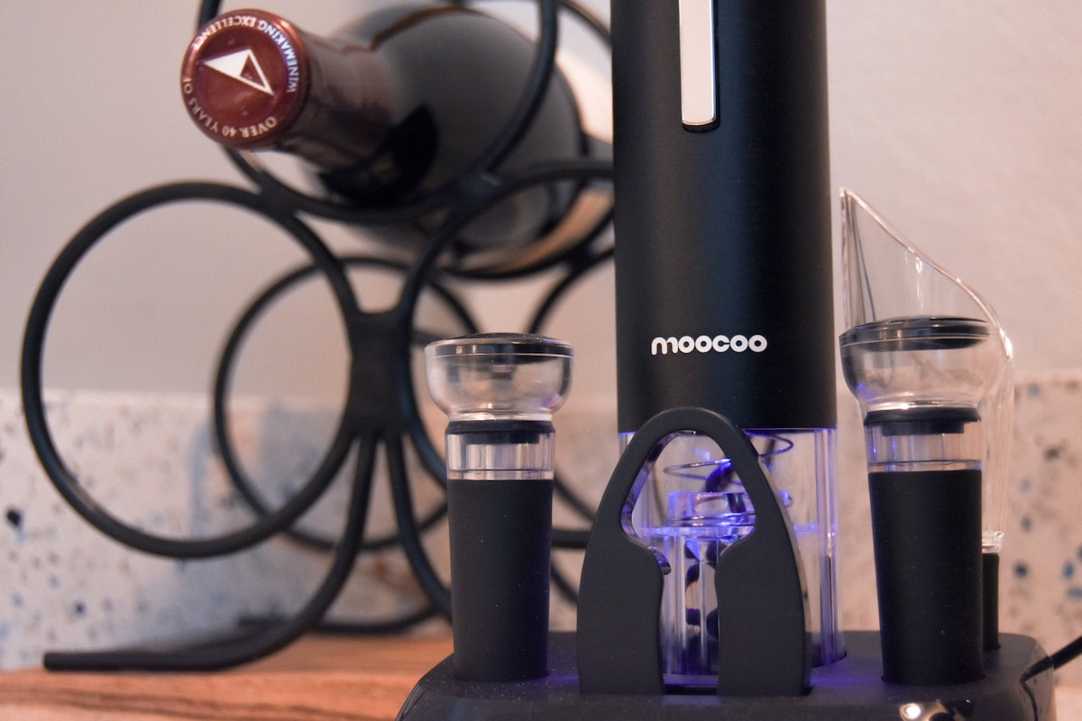 An electric wine opener is displayed on a countertop, accompanied by two wine glasses. A wine bottle is positioned in a decorative metal rack in the background, adding an element of sophistication to the setting.