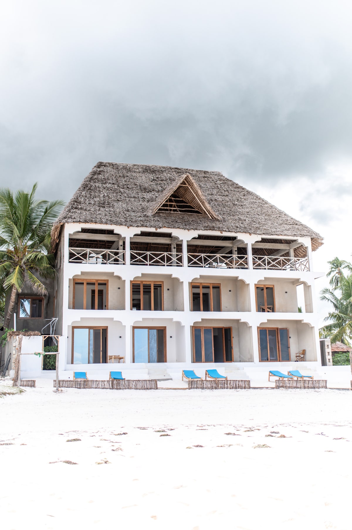 A three-story beachfront building features a thatched roof and multiple large glass doors. Sun loungers are placed along the sandy area in front. Lush palm trees are visible beside the structure, contributing to the tropical ambiance.