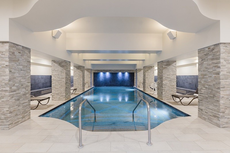 An indoor swimming pool is depicted with a calming blue water surface. Soft lighting highlights the sleek stone columns and tiled walls. Lounge chairs line the edges, inviting relaxation in a serene environment.
