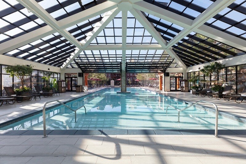 An indoor swimming pool is housed under a large glass roof, allowing natural light to fill the space. Lounge chairs are positioned around the pool, and potted plants are placed along the edges, creating a serene atmosphere for relaxation.