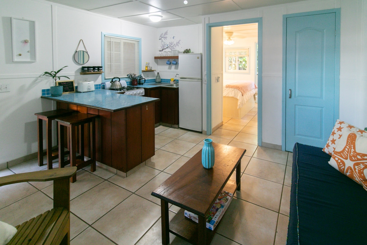 A well-equipped kitchen area is visible, featuring modern appliances and a blue tiled countertop. Adjacent seating is provided at a wooden bar, with a cozy living space that includes a dark sofa and a coffee table. Soft light enters through a nearby door leading to the bedroom.