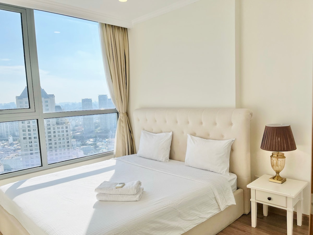 A spacious bedroom features a comfortable bed with a plush headboard. Natural light floods the room through large windows, offering a view of the city skyline. Soft curtains frame the windows, while a bedside table with a lamp complements the neutral decor.