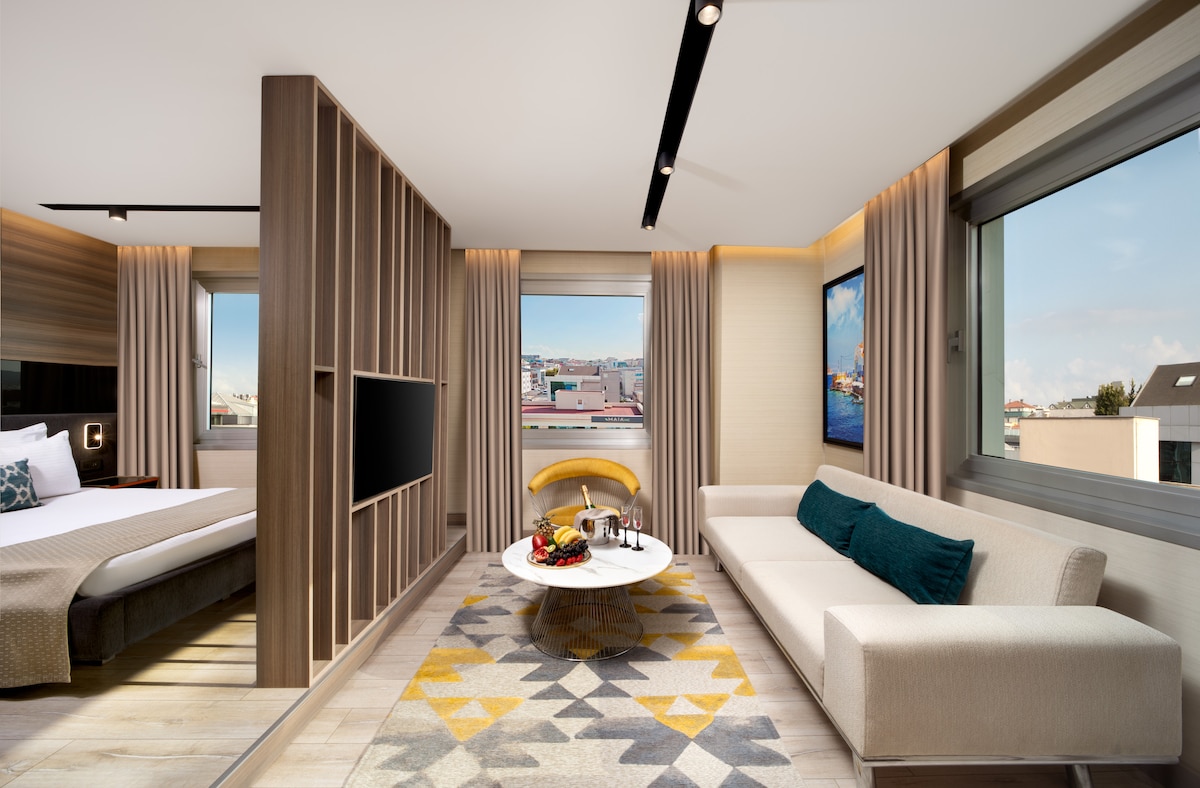 A modern living area features a comfortable beige sofa and a round coffee table adorned with fresh fruit. Large windows allow natural light to fill the space, showcasing city views. A cozy sleeping area is separated by a decorative partition with a television mounted on the wall.