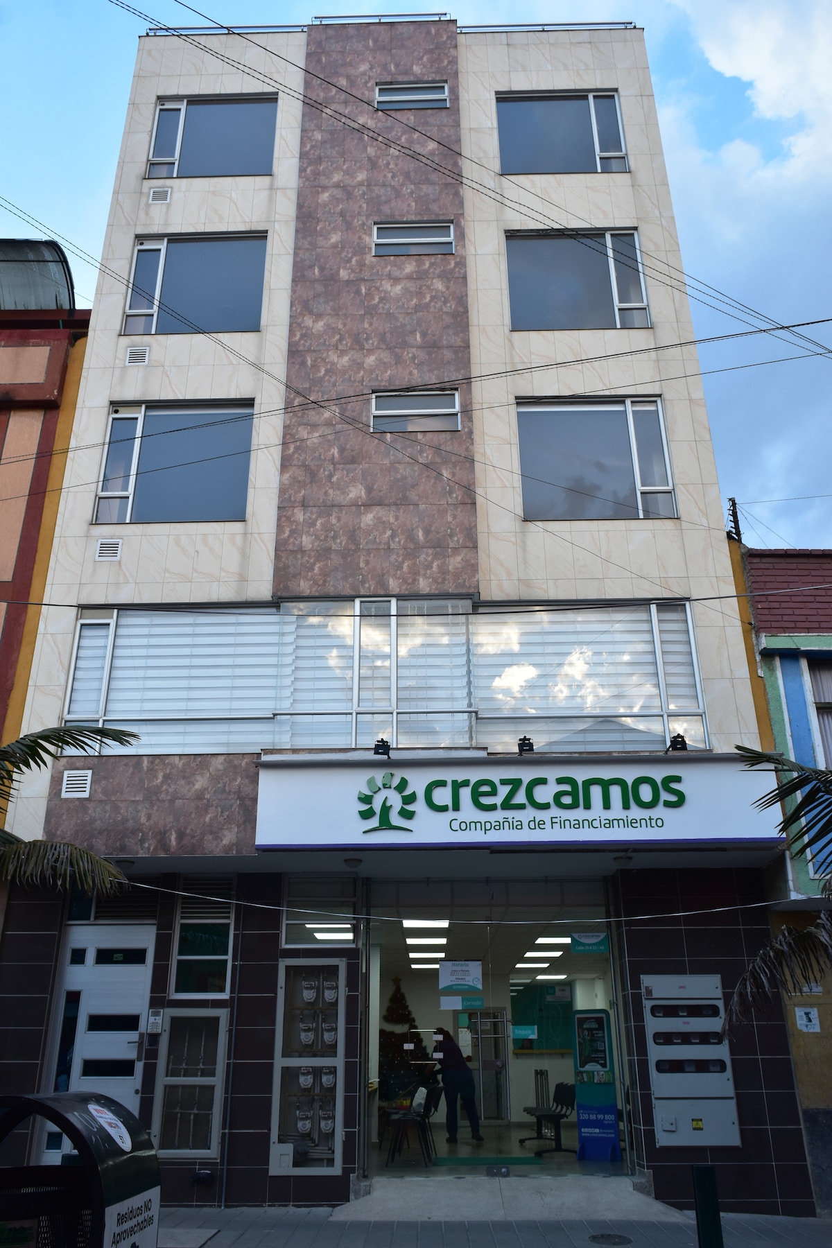 The building features a modern façade with a combination of light-colored tiles and dark stone accents. Large windows provide natural light to the interior. A commercial space is located on the ground floor, displaying signage for 'crezcamos' with visitors visible inside.