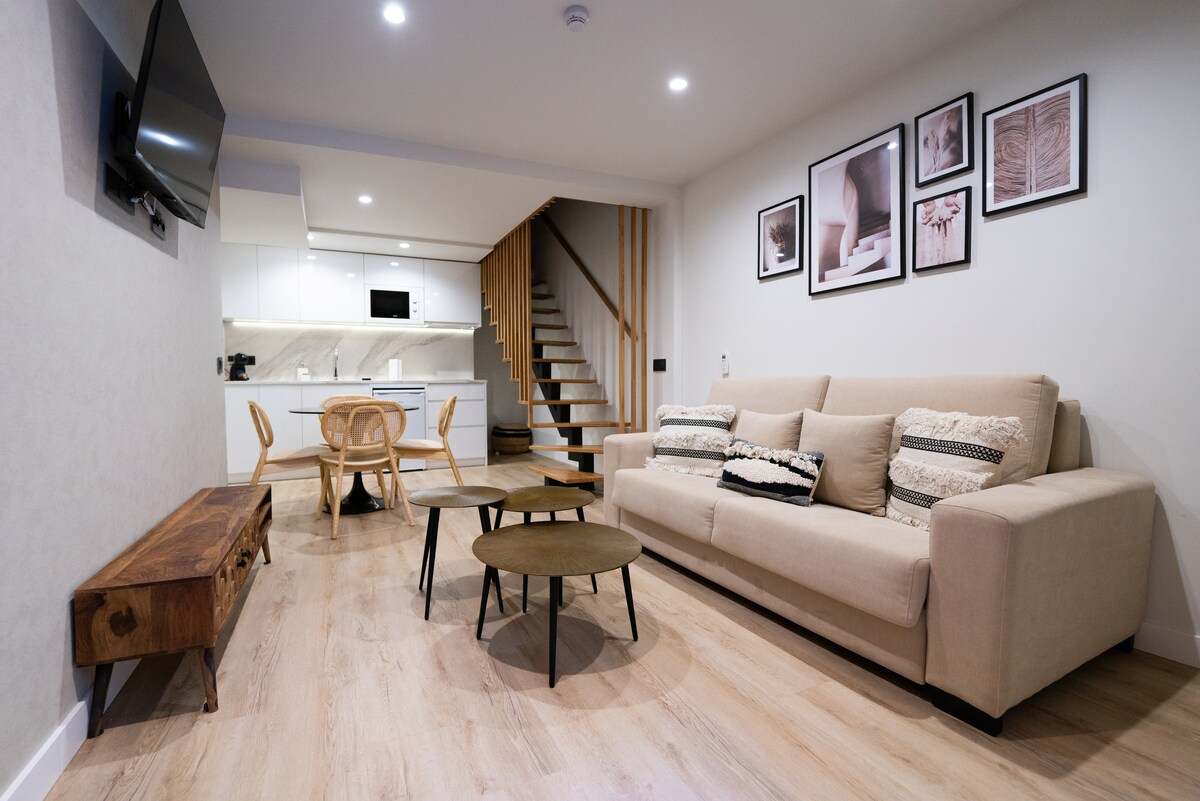 A modern living area is displayed, featuring a beige sofa adorned with patterned cushions. A set of three coffee tables in varying sizes is positioned in front of the sofa. A television is mounted on the wall, while a staircase leads to the upper level. Light wood flooring complements the overall design.