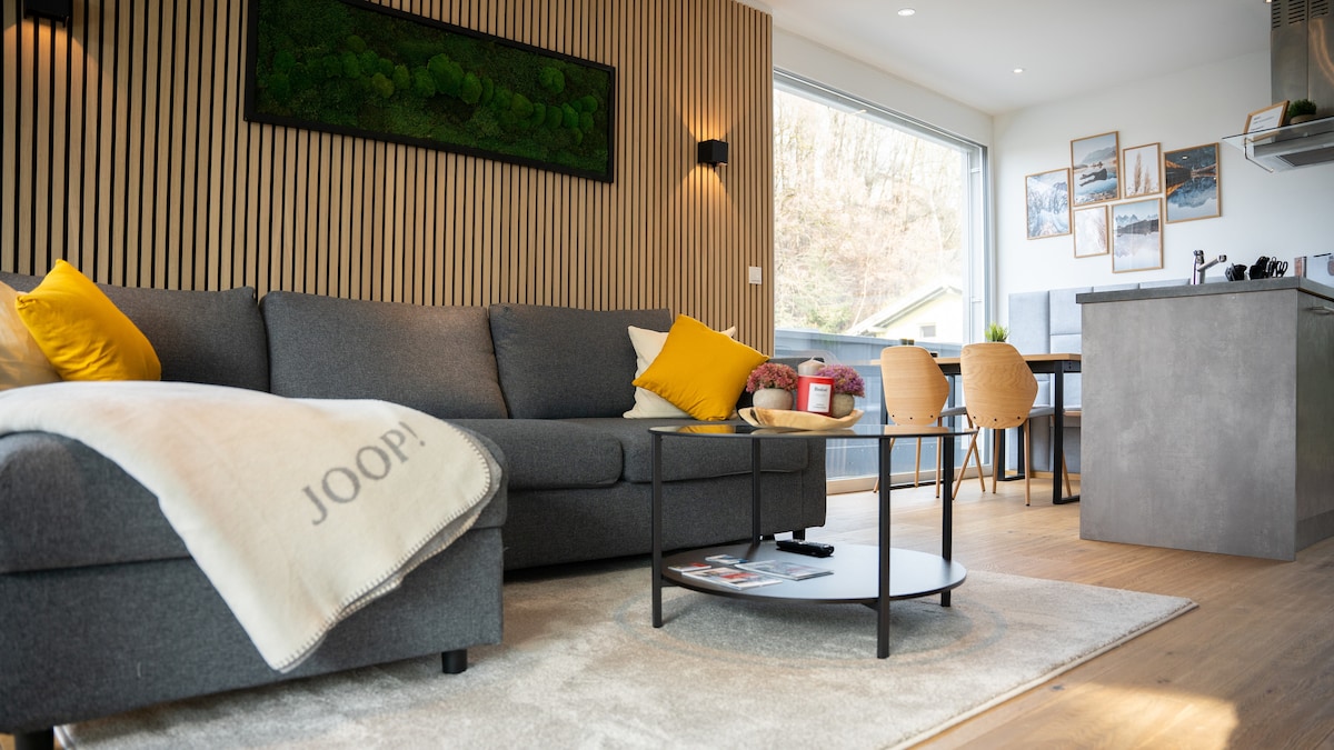 A spacious living area is presented featuring a gray sectional sofa adorned with a yellow pillow. A soft blanket is draped over the side. A low coffee table sits on a light rug, with a sleek dining table and chairs visible in the background, illuminated by natural light.