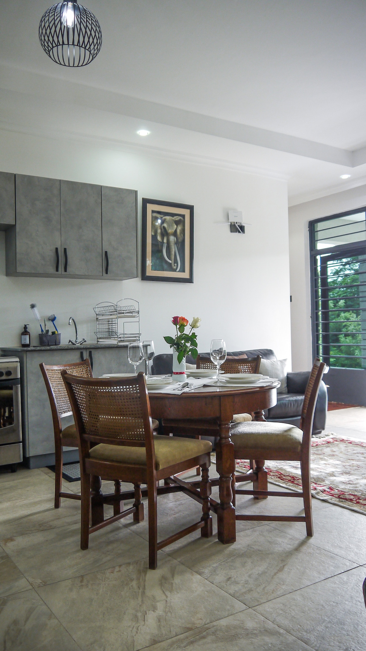 A dining area is visible, featuring a round wooden table surrounded by four classic chairs. A small vase with flowers sits at the center of the table. The kitchenette is equipped with gray cabinetry and essential appliances, while a cozy seating area is positioned nearby, offering a view through large windows.