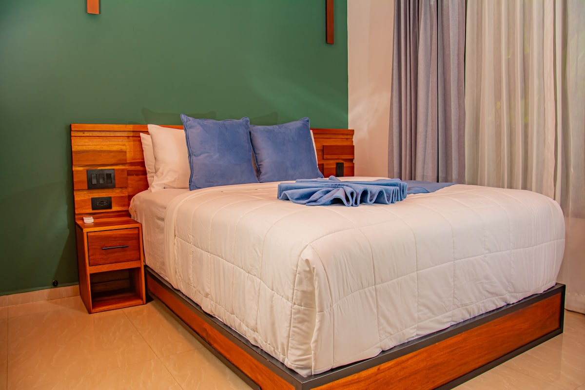 A spacious bed with a crisp white quilt and two blue pillows is positioned against a green accent wall. Natural light filters through tall, light-colored curtains, providing a serene ambiance. A wooden nightstand is visible beside the bed, enhancing the room's modern design.