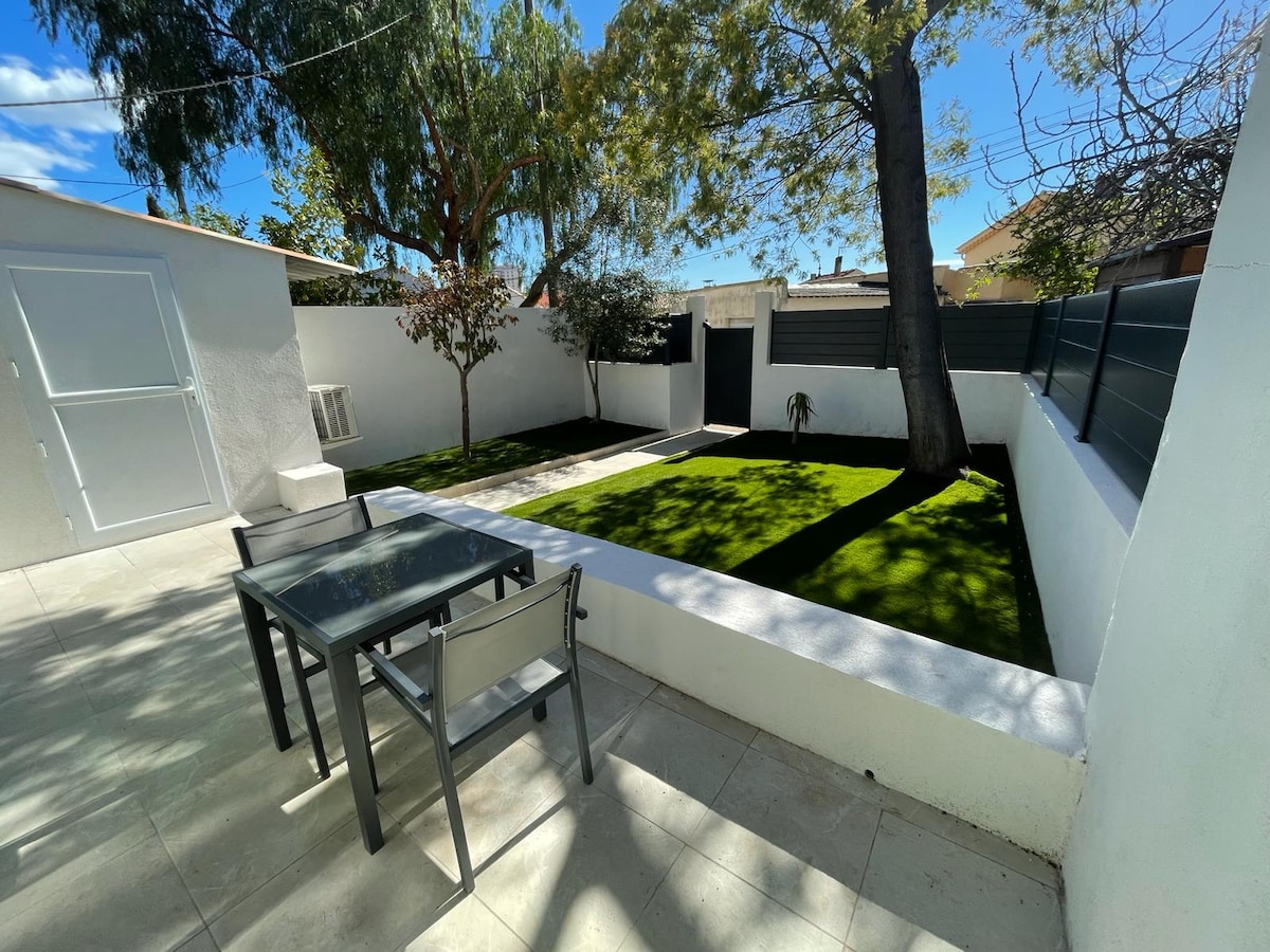 An outdoor space features a paved patio with a glass table and four chairs, surrounded by a neatly maintained garden area. The space is complemented by greenery, including a mature tree, providing shade and a serene setting.