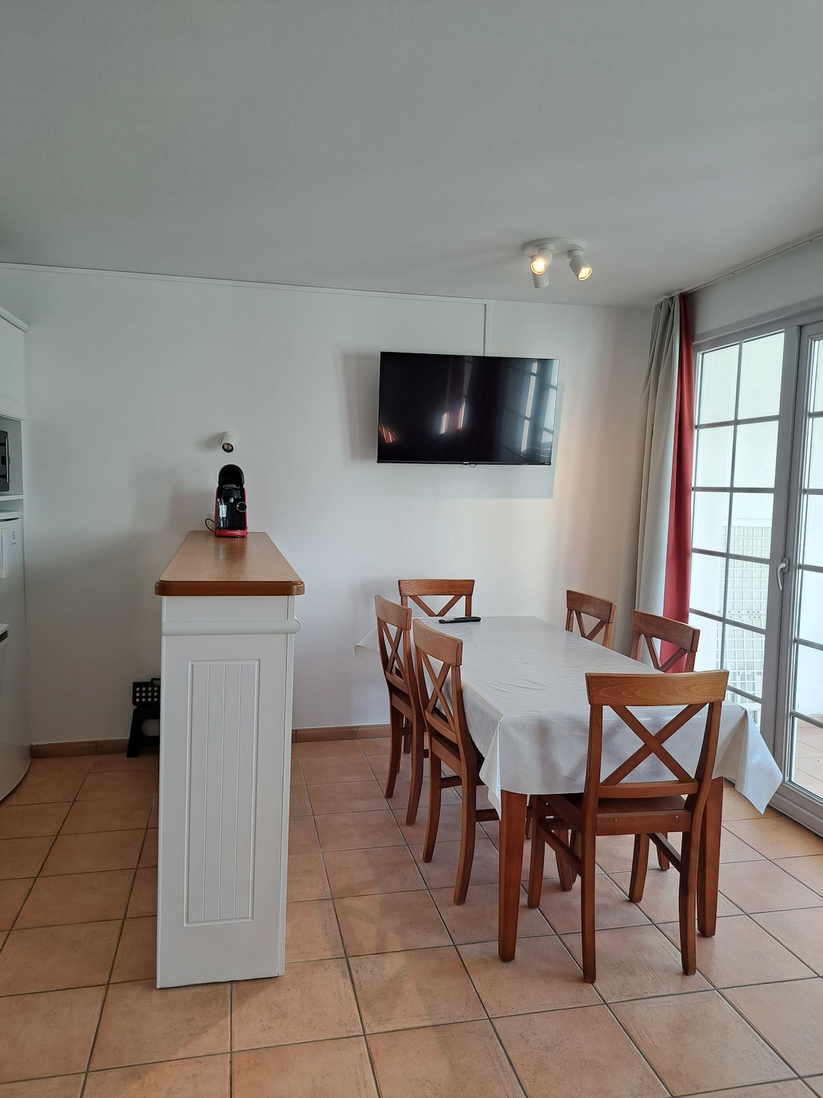 The dining area features a rectangular table with seating for six, positioned under a wall-mounted television. Light-colored cabinetry and a small counter complement the space, while natural light filters through the nearby glass doors leading to the terrace.