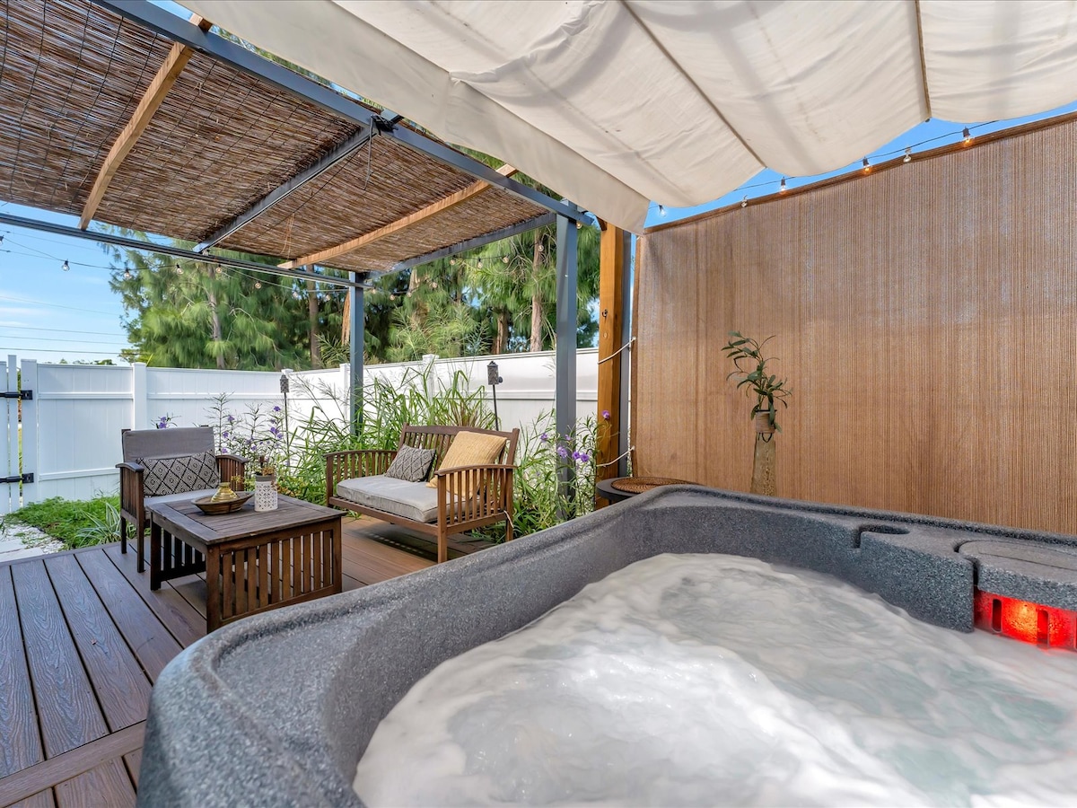 A sheltered hot tub area features flowing water and relaxation seating. A wooden table with light decor complements the surrounding greenery. Soft drapes filter sunlight, creating a serene atmosphere for unwinding.