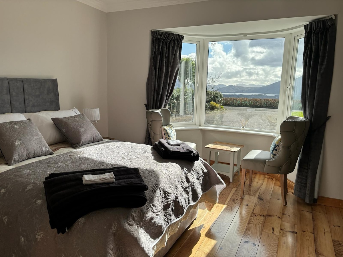 A bright bedroom features a spacious bed adorned with light-colored bedding and neatly arranged black towels. Two armchairs sit beside a small table, positioned under a large bay window that frames panoramic views of the surrounding landscape.