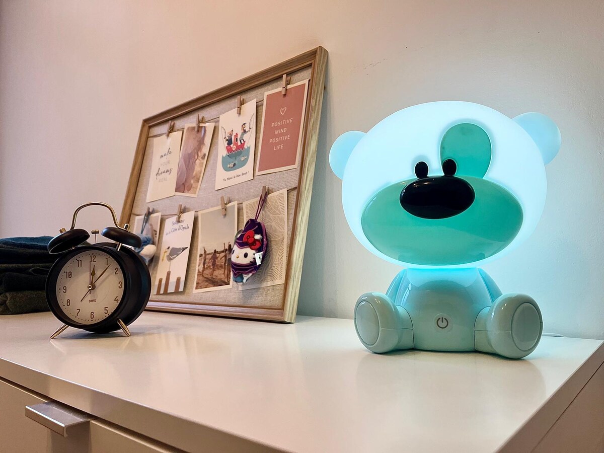 A playful bedside table features a blue bear-shaped lamp glowing softly, providing gentle illumination. An analog clock displays a classic design alongside a framed collage of photos and notes, pinned with clips. Additional personal items are neatly arranged, enhancing the cozy atmosphere.