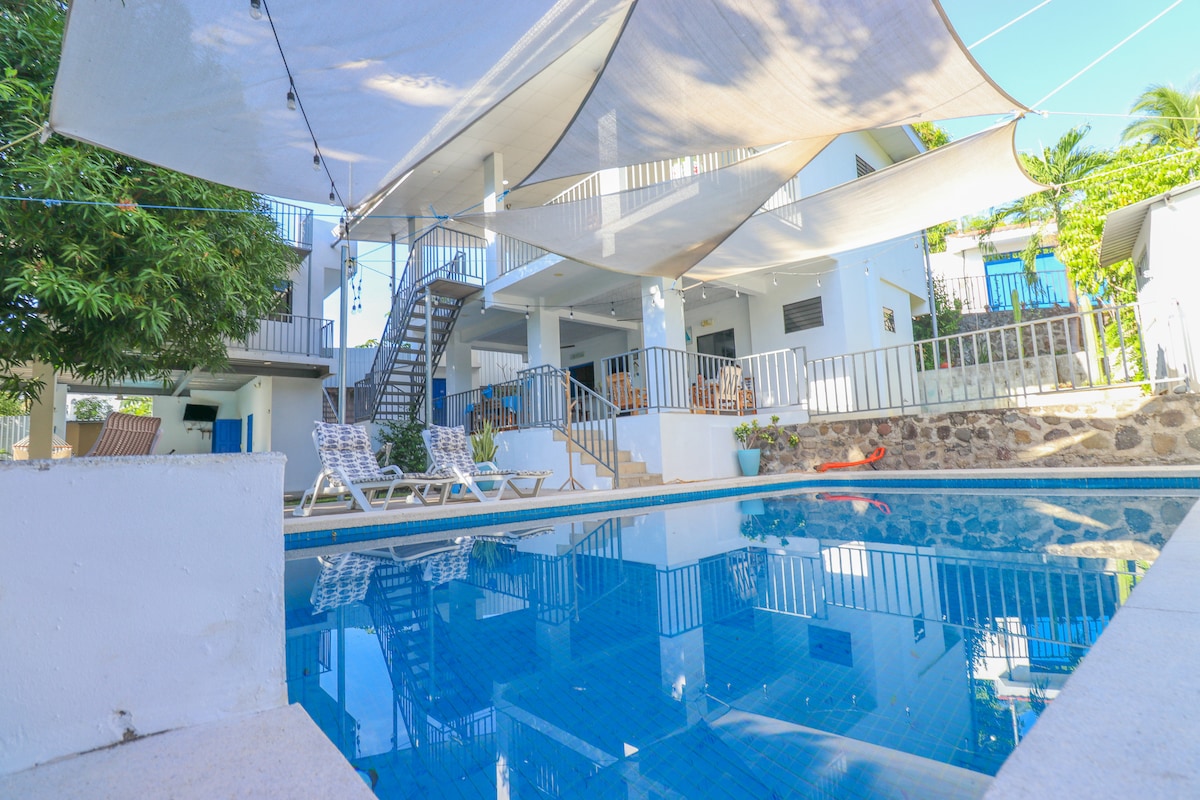 A private pool is seen with a clear blue surface, surrounded by a spacious deck featuring lounge chairs. Shade is provided by large fabric canopies overhead, while a staircase is visible, leading to a second level of the property. Lush greenery complements the serene setting.
