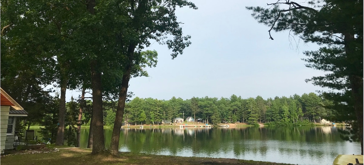 A serene view of the tranquil lake is framed by tall trees. The calm water reflects the surrounding lush greenery and distant houses. A natural shoreline creates an inviting atmosphere for outdoor relaxation.