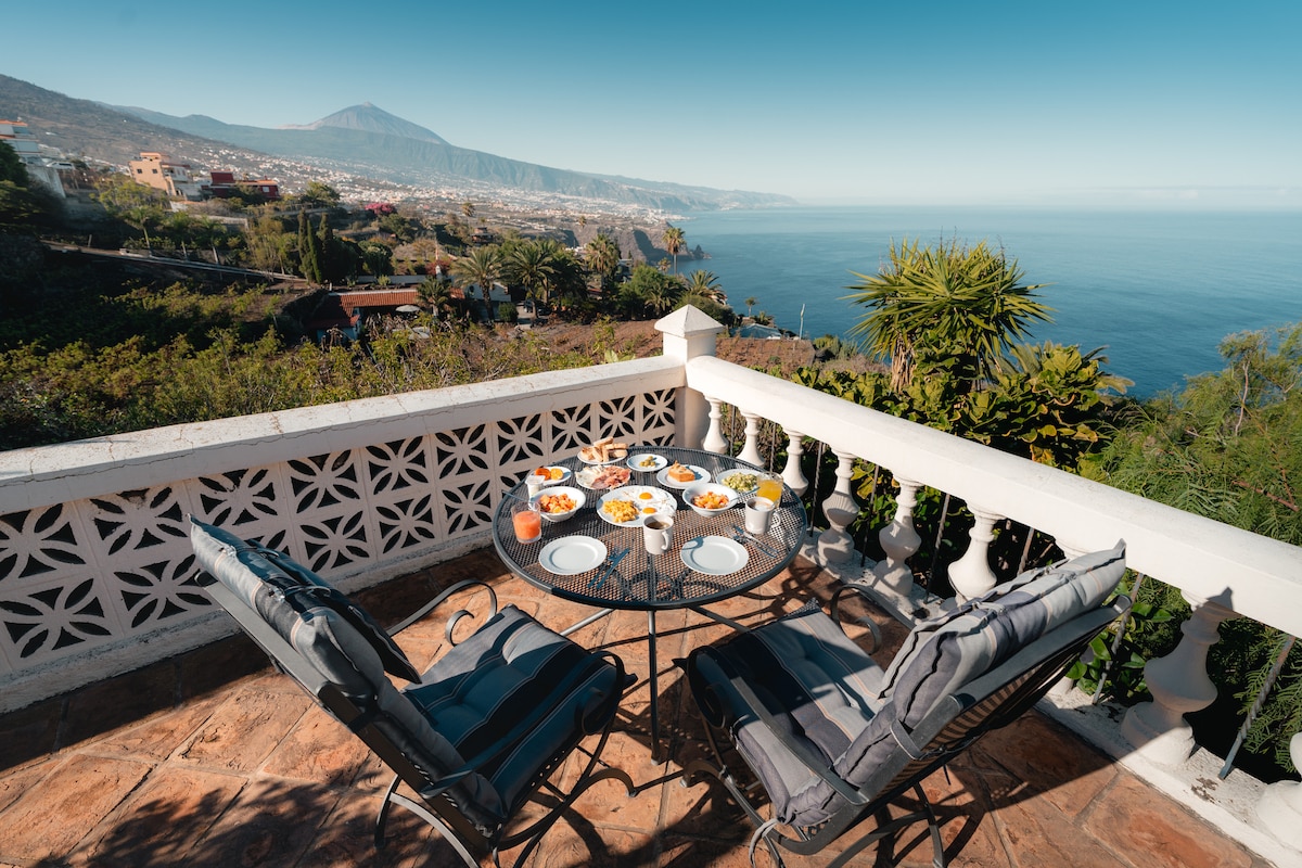 A terrace dining area is set with a round table, adorned with a selection of fruits and dishes. Comfortable seating is provided by cushioned chairs, with a view overlooking the ocean and distant mountains, creating a serene outdoor space.
