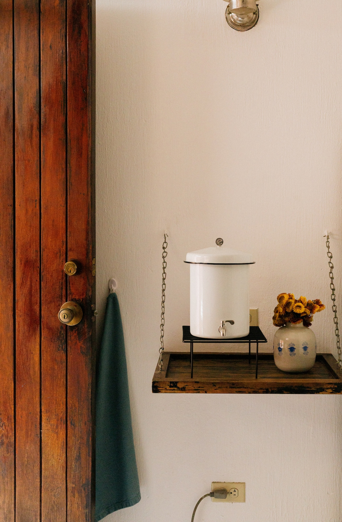 A wooden door with a brass handle opens into a small entryway. A white water dispenser is placed on a wooden shelf supported by chains, alongside a ceramic vase containing dried flowers. A towel hangs from a hook nearby.