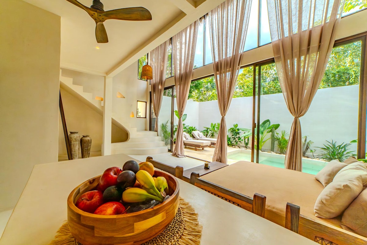 An inviting open-plan living space features large windows draped with light curtains, allowing natural light to fill the area. A decorative wooden bowl filled with fresh fruit is placed on a neutral-toned table. The view extends to the private pool and lush greenery outside.