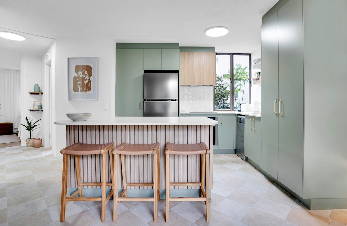 A modern kitchen area features sleek cabinetry in soft green and natural wood tones. A central island with bar seating for three is complemented by a contrasting white countertop. Large windows allow natural light to brighten the space, enhancing the inviting atmosphere.