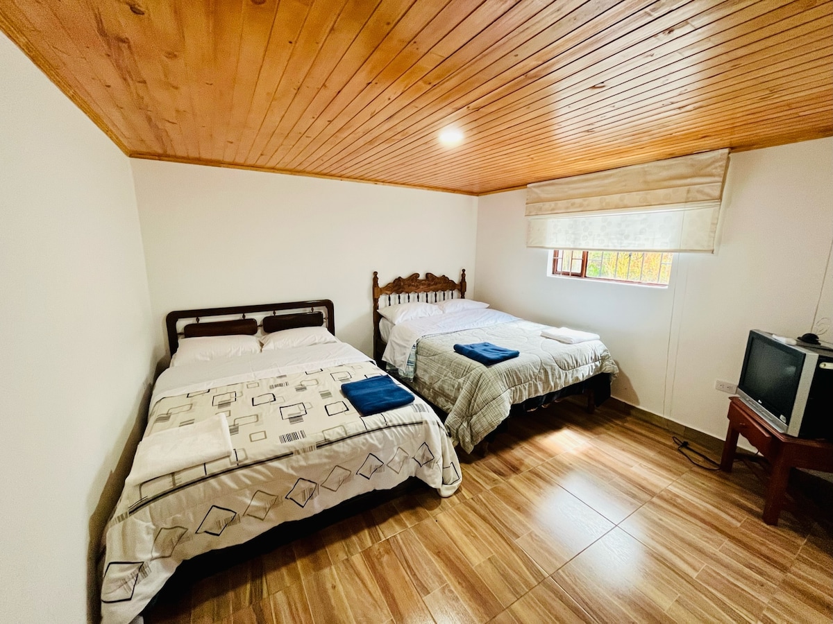 A spacious room features two beds, each covered with neutral bedding and neatly arranged towels. Natural light enters through a window dressed with soft curtains. A wooden ceiling and tile flooring create a calming environment, accompanied by a television positioned on a side table.