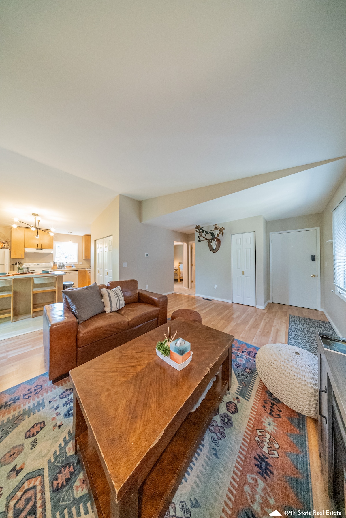 The living area features a leather sofa positioned next to a wooden coffee table adorned with a potted plant. A patterned rug covers the floor, and light enters through a nearby window. Open access leads to a well-lit kitchen area in the background.
