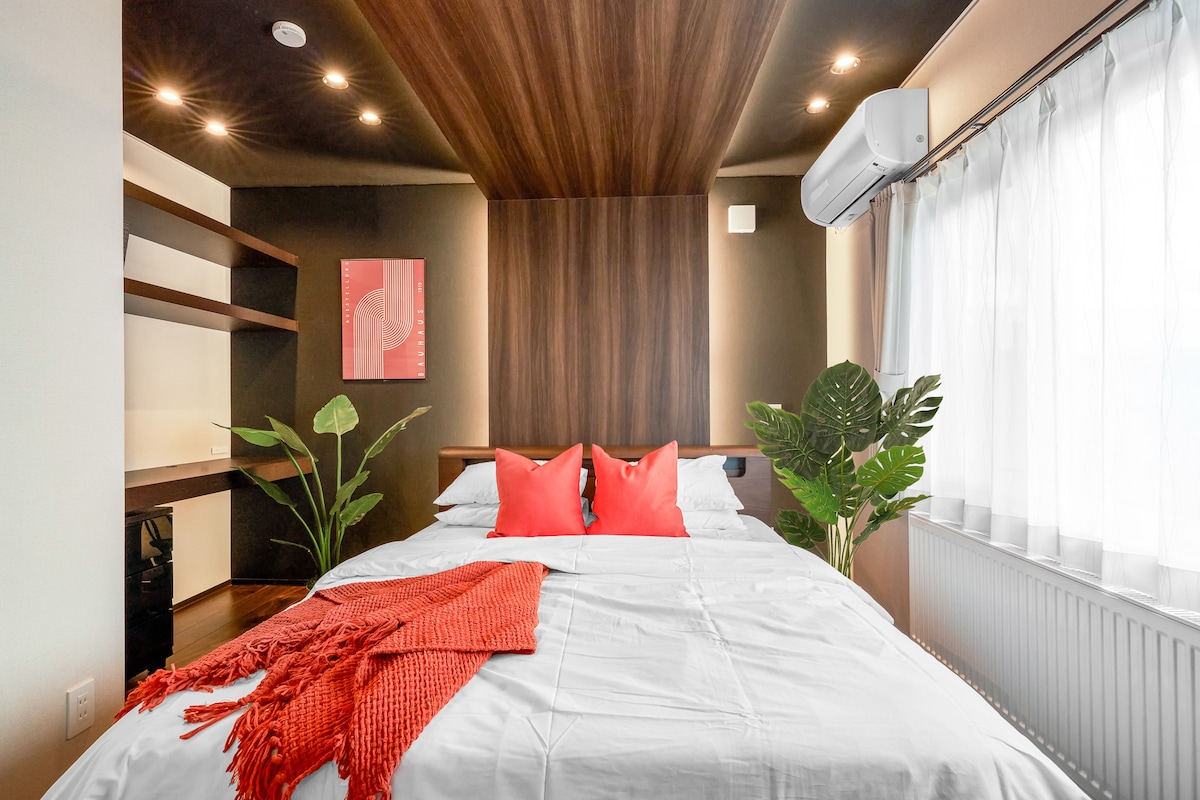 A spacious bedroom features a large bed adorned with red accent pillows. A warm wooden ceiling contrasts with a modern dark wall backdrop. Natural light filters through large sheer curtains, while a green plant and minimalist shelving add a touch of vibrancy to the space.