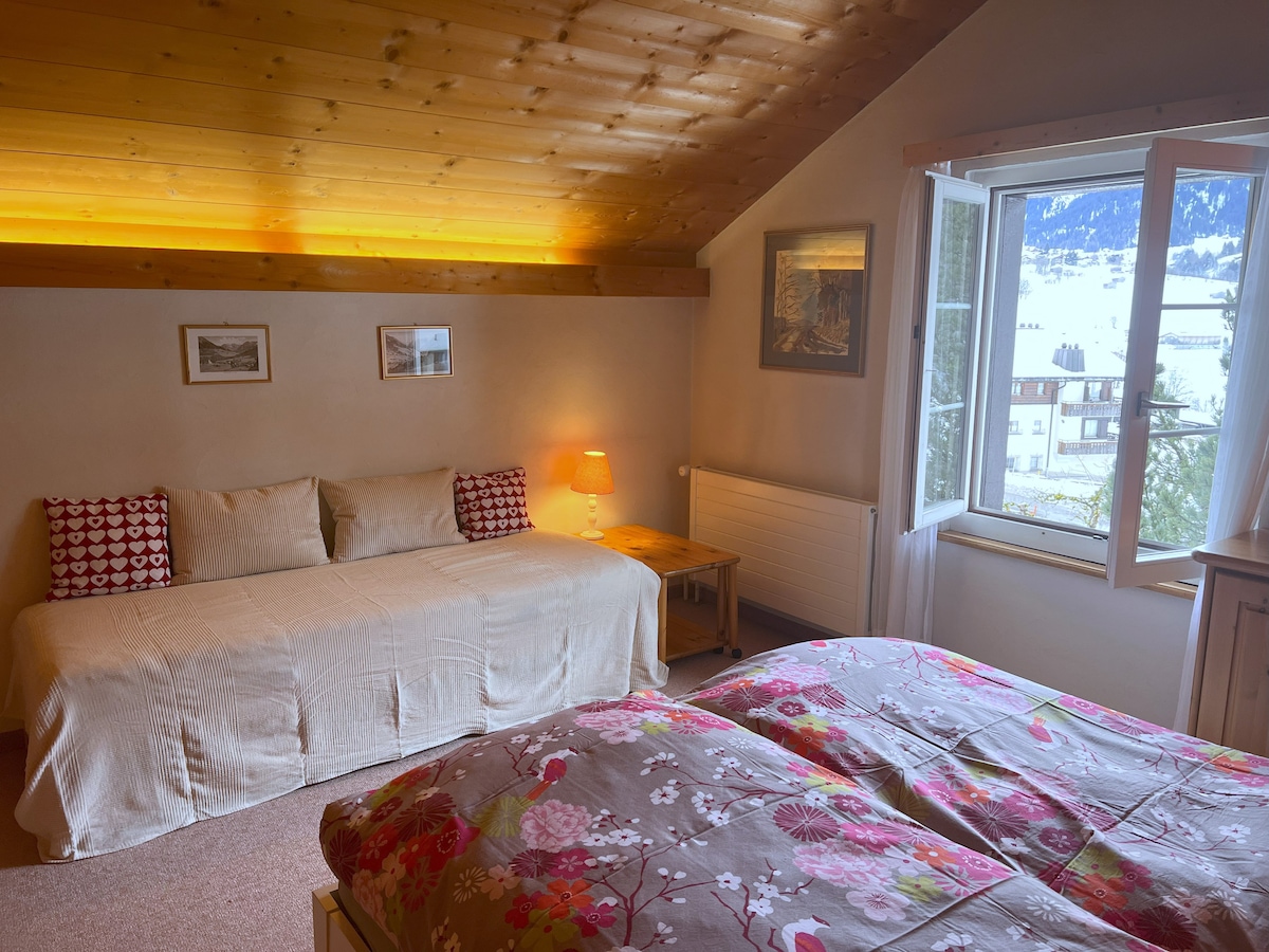 A spacious bedroom is featured, showcasing a comfortable double bed and a day bed that can be separated into two beds. Natural light enters through a window, which opens to beautiful mountain views. Warm wood paneling highlights the cozy atmosphere.
