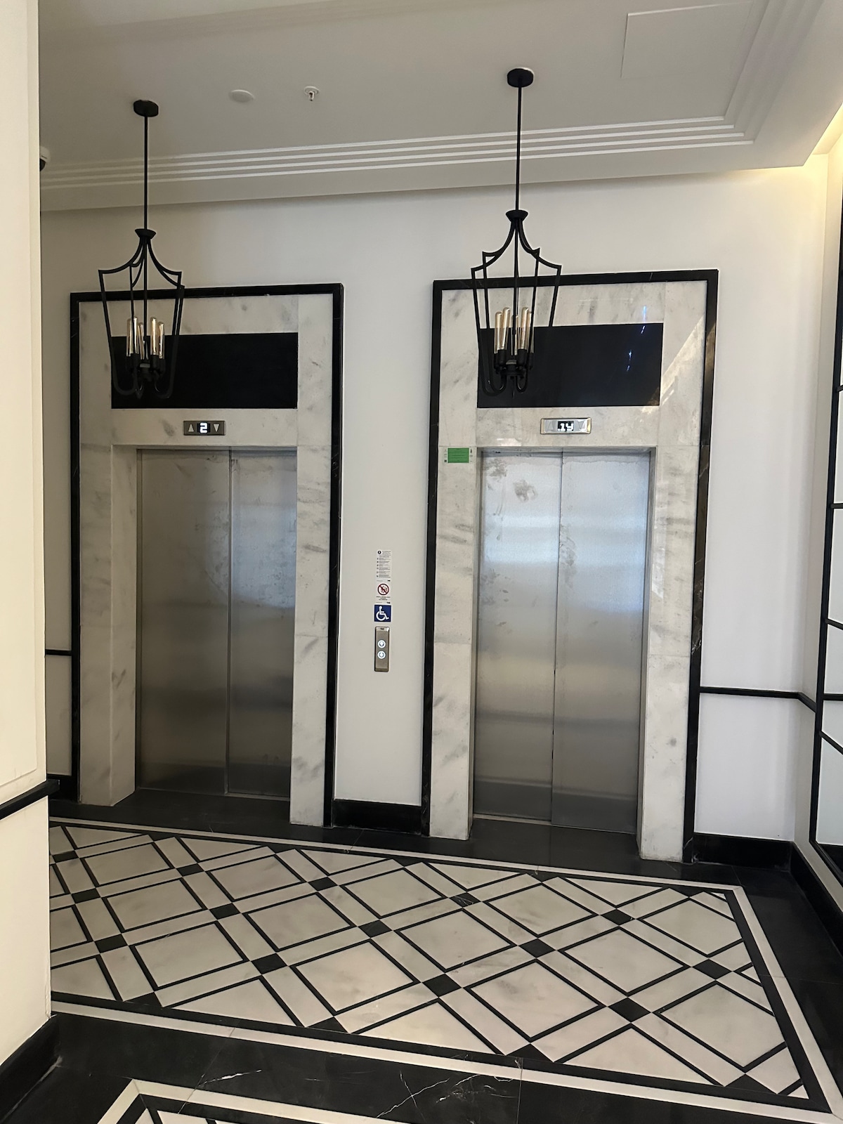 Two modern elevators are positioned side by side, framed by elegant marble walls in a stylish lobby. Luxurious light fixtures hang overhead, while a geometric patterned floor adds sophistication to the space.