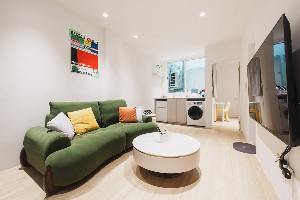 A modern living space features a green sectional sofa adorned with yellow and black cushions. A circular coffee table sits in front of it. A large TV is mounted on the wall. A window reveals a glimpse of the kitchen area, which includes a washing machine.