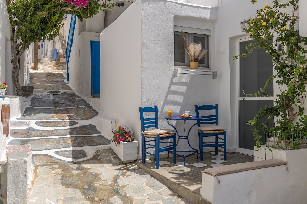 A charming outdoor seating area is positioned near a whitewashed building. A small round table is accompanied by two blue chairs, with a glass of orange juice placed on the table. Lush greenery and colorful flowers enhance the inviting entrance, with cobblestone paths leading away.
