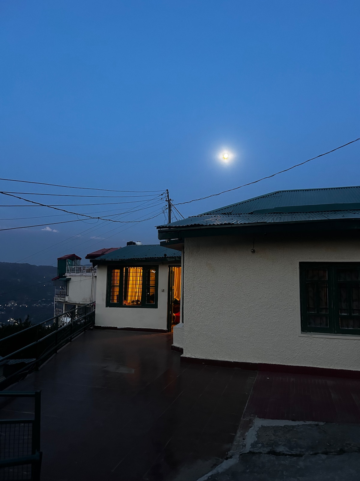 An outdoor view of the cottage at twilight, featuring a clear sky and a visible moon. The space highlights a balcony area, with warm light spilling from windows. A hilly landscape is faintly seen in the background, surrounded by electrical wires.